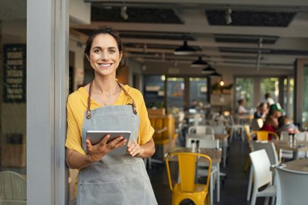 Portrait of happy woman standing at doorway of her store. Cheerful mature waitress waiting for clients at coffee shop. Successful small business owner in casual wearing grey apron standing at entrance